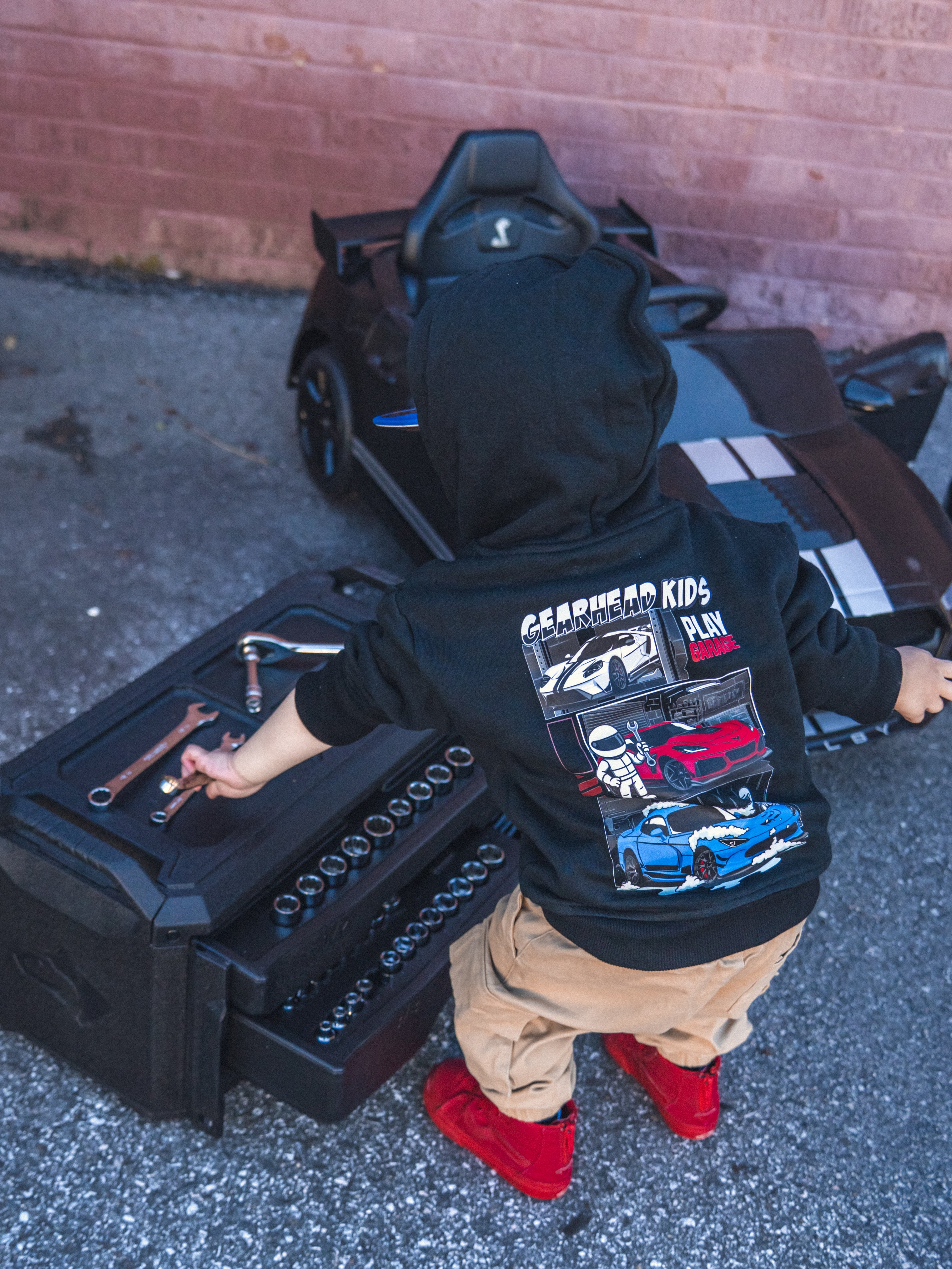 Child playing with a powerwheels car, wearing a Gearhead Kids American Muscle Car black hoodie featuring Ford GT, Corvette and Viper muscle cars.  Car themed clothing for kids. 