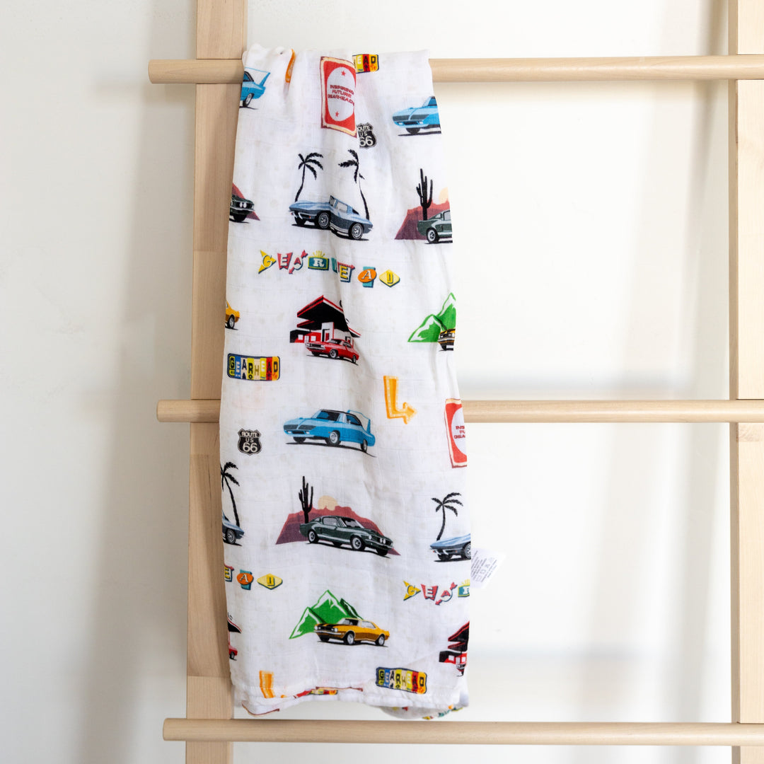 A muslin bamboo swaddle blanket with a pattern of classic american muscle cars displayed on a wooden ladder.