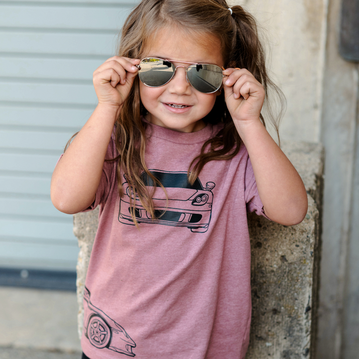 Young girl wearing a pink t-shirt with Porsche Carrera GT graphics, standing against a wall - Gearhead Kids