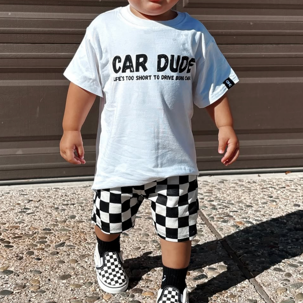 Child wearing a white t-shirt with 'CAR DUDE' text, checkered shorts, and matching shoes on a pavement.
