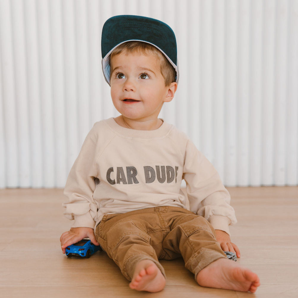 A little child sitting on the floor wearing a beige sweatshirt with the text 'CAR DUDE' on it, paired with brown pants and a green hat - Gearhead Kids