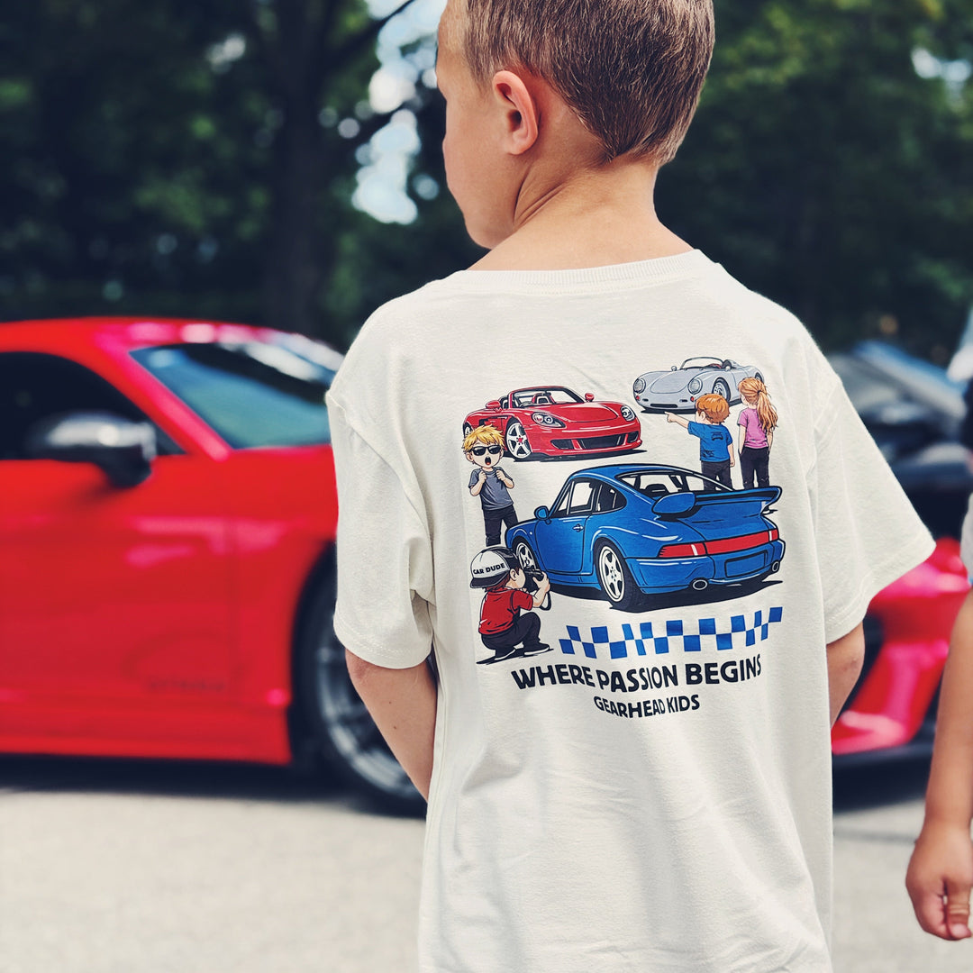 A child wearing an off-white t-shirt with a graphic featuring classic sports cars 964 Turbo, 550 Spyder and Mid-engine V10 Sports car and the text 'WHERE PASSION BEGINS | GEARHEAD KIDS'. The background shows a red sports car and green trees.