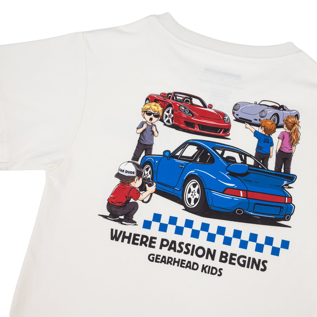 A close-up of an off-white short-sleeve t-shirt featuring a colorful cartoon graphic of children with mid-engine V10 supercar, a 964 turbo, and a classic 550 spyder sports cars and the text 'WHERE PASSION BEGINS GEARHEAD KIDS' 
