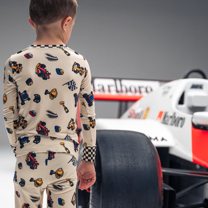 Little boy standing next to a Formula 1 car wearing formula 1 themed pajamas featuring race cars, trophies, champagne and checkered flags.  | Gearhead Kids