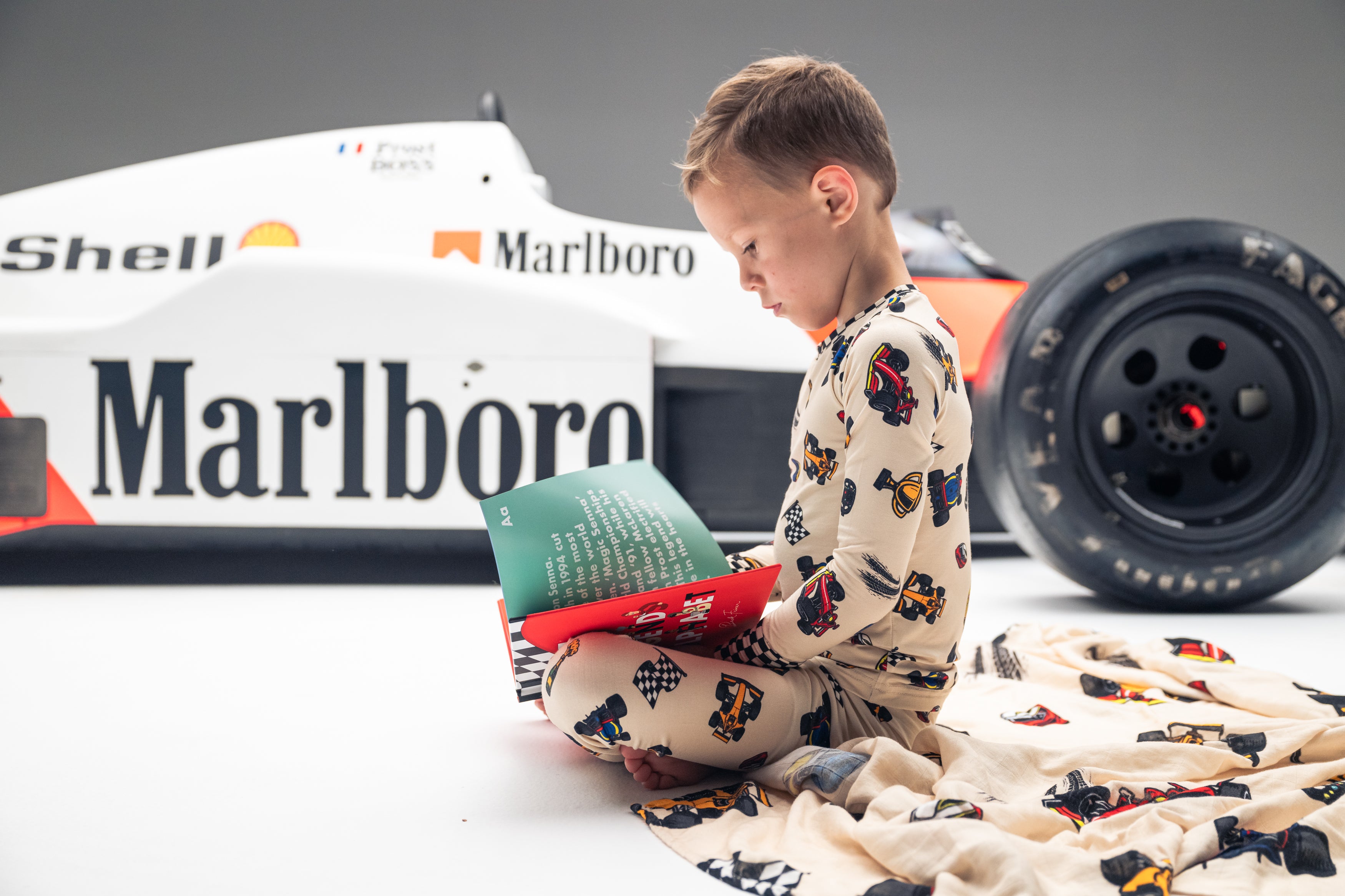 Little boy sitting next to a Formula 1 race car in Grand Prix themed bamboo pajamas reading the F1 Legends Alphabet Book | Gearhead Kids