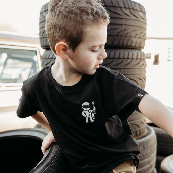 Boy Wearing a Gearhead Kids - Play Garage t-Shirt surrounded by tires.  Car Apparel For Kids.  