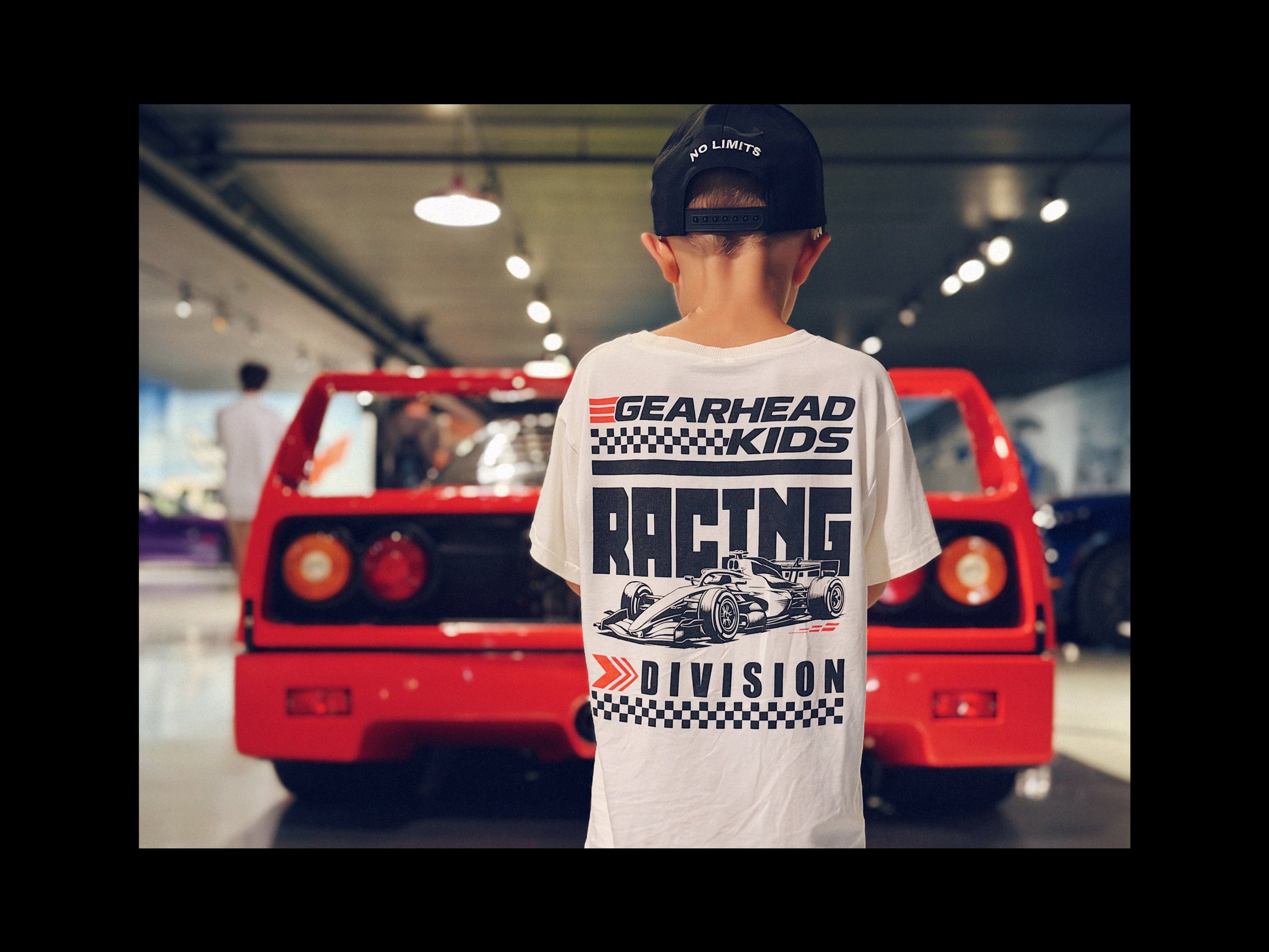 a boy wearing Gearhad Kids Racing Division white t-shirt with a open wheel race car graphic and bold Racing Division text standing in front of a Race Car
