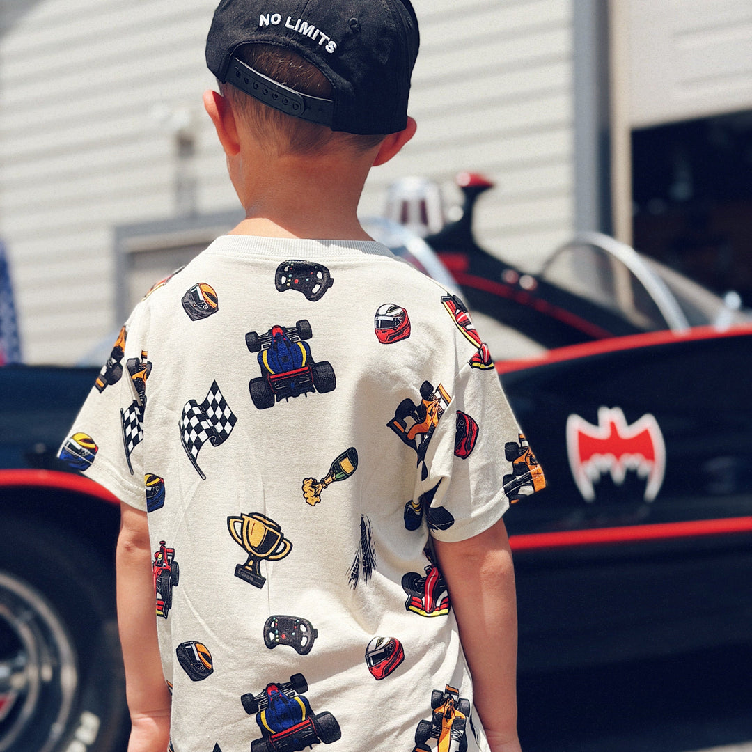 A child viewed from behind wearing a Gearhead Kids t-shirt with racing-themed print (race cars, checkered flags, trophies) and a black baseball cap with 'NO LIMITS' on the back, standing in front of a red and black race car. 