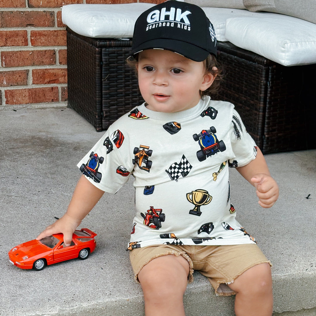 A toddler wearing a race car themed shirt with Grand Prix winning cars and trophies and a black GHK hat Gearhead Kids.  The boy is holding a small red car in his hand.
