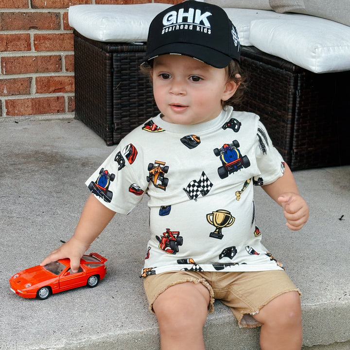 A toddler wearing a race car themed shirt with Grand Prix winning cars and trophies and a black GHK hat Gearhead Kids.  The boy is holding a small red car in his hand.