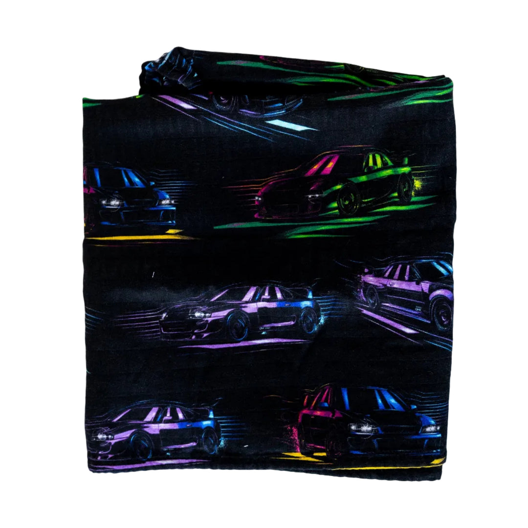A folded swaddle blanket with a pattern of colorful, glowing images of classic japanese cars on a dark background - Gearhead Kids