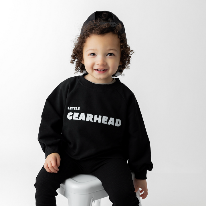 A toddler wearing a black oversized 'LITTLE GEARHEAD' sweatshirt and a cap, sitting on a white stool against a white background. - gearhead kids