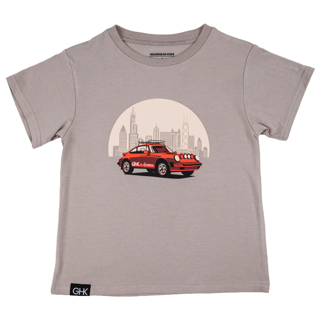 A kids' short-sleeve crewneck t-shirt in mocha color, featuring a graphic print of a red vintage safari 911 car in front of Chicago skyline. The shirt has a black 'GHK' brand label on the bottom left hem.