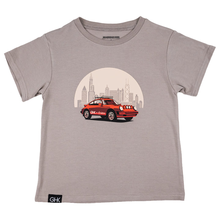 A kids' short-sleeve crewneck t-shirt in mocha color, featuring a graphic print of a red vintage safari 911 car in front of Chicago skyline. The shirt has a black 'GHK' brand label on the bottom left hem.