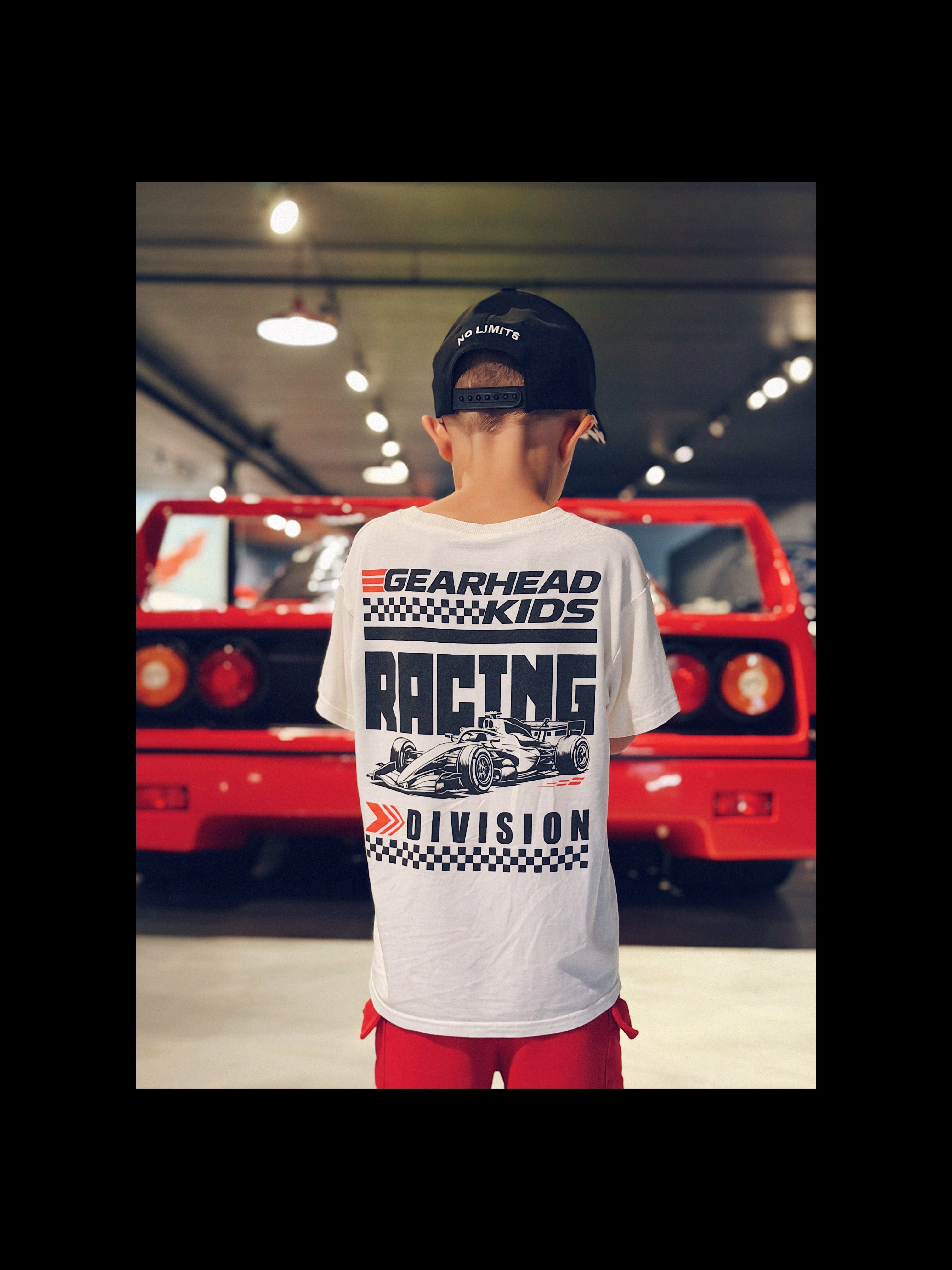 a boy wearing Gearhad Kids Racing Division white t-shirt with a open wheel race car graphic and bold Racing Division text standing in front of a Race Car