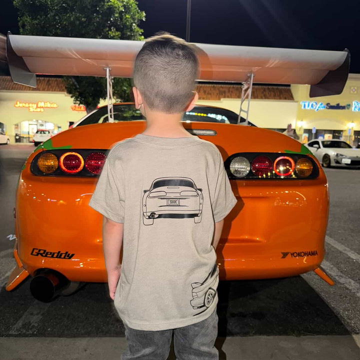Little boy wearing a Toyota Supra MK4 t-shirt in Stone color - standing in front of a Toyota Supra Orange car.  | Gearhead Kids