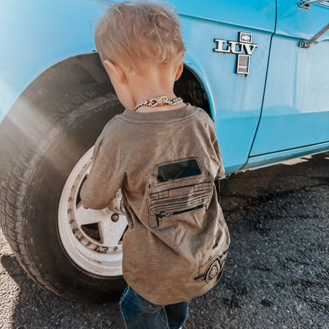 Little boy wearing a VW GTI T-shirt in olive standing next to a blue car. 