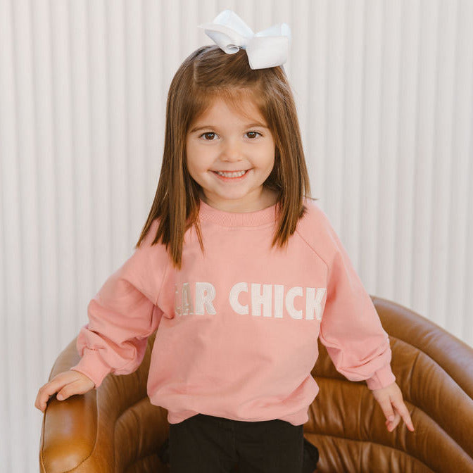 A young girl wearing a pink sweatshirt with the text 'CAR CHICK' on it, sitting in a brown chair with a white background. - Gearhead Kids